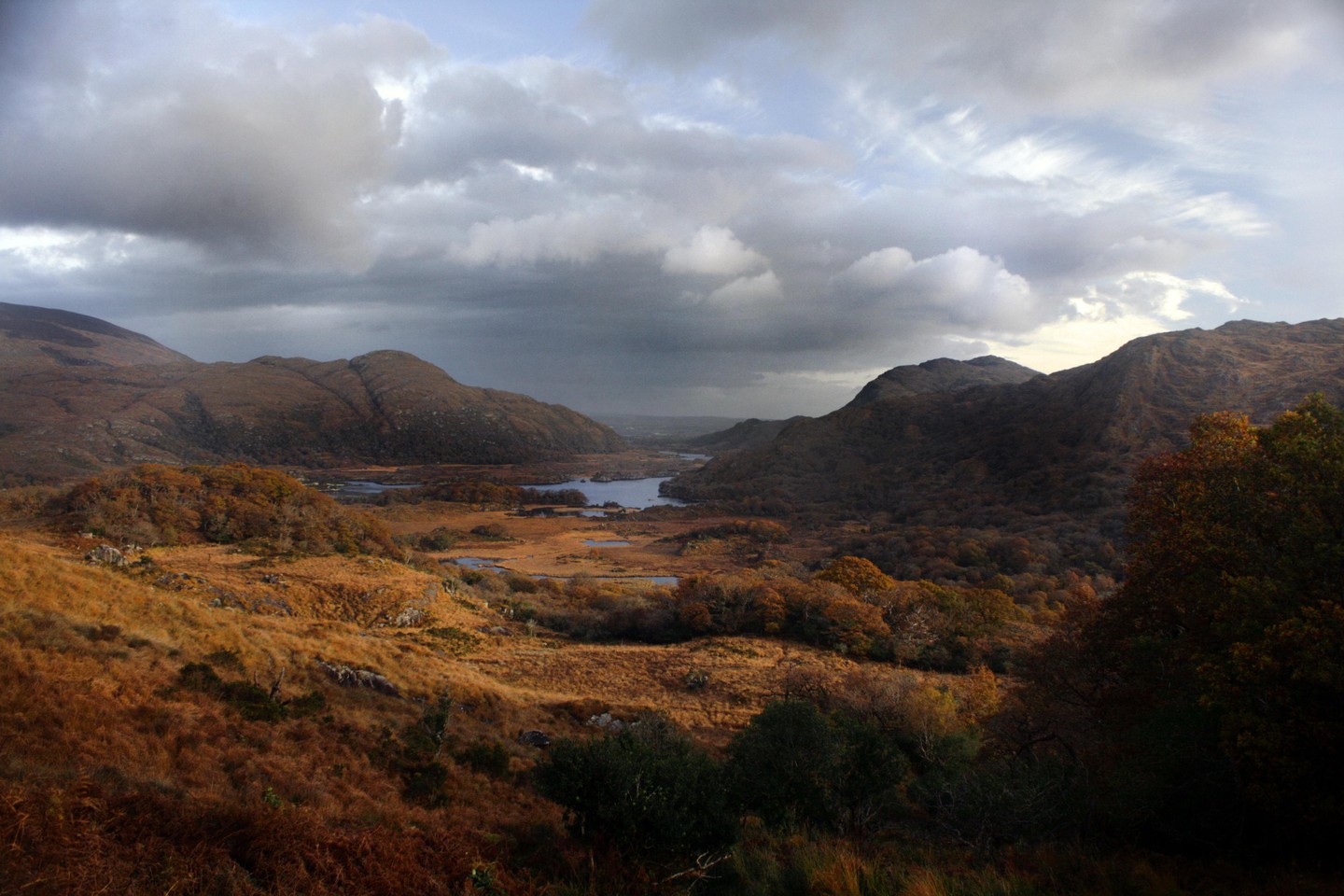 A valley with a river and lakes is surrounded by mountains under a cloudy sky.