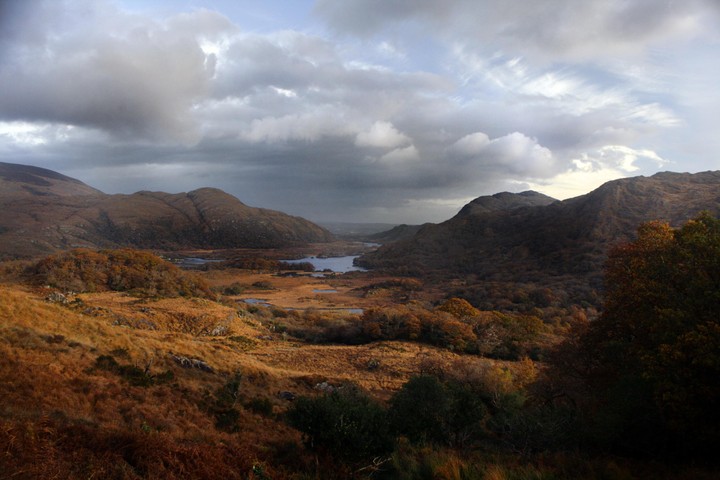 A valley with a river and lakes is surrounded by mountains under a cloudy sky.