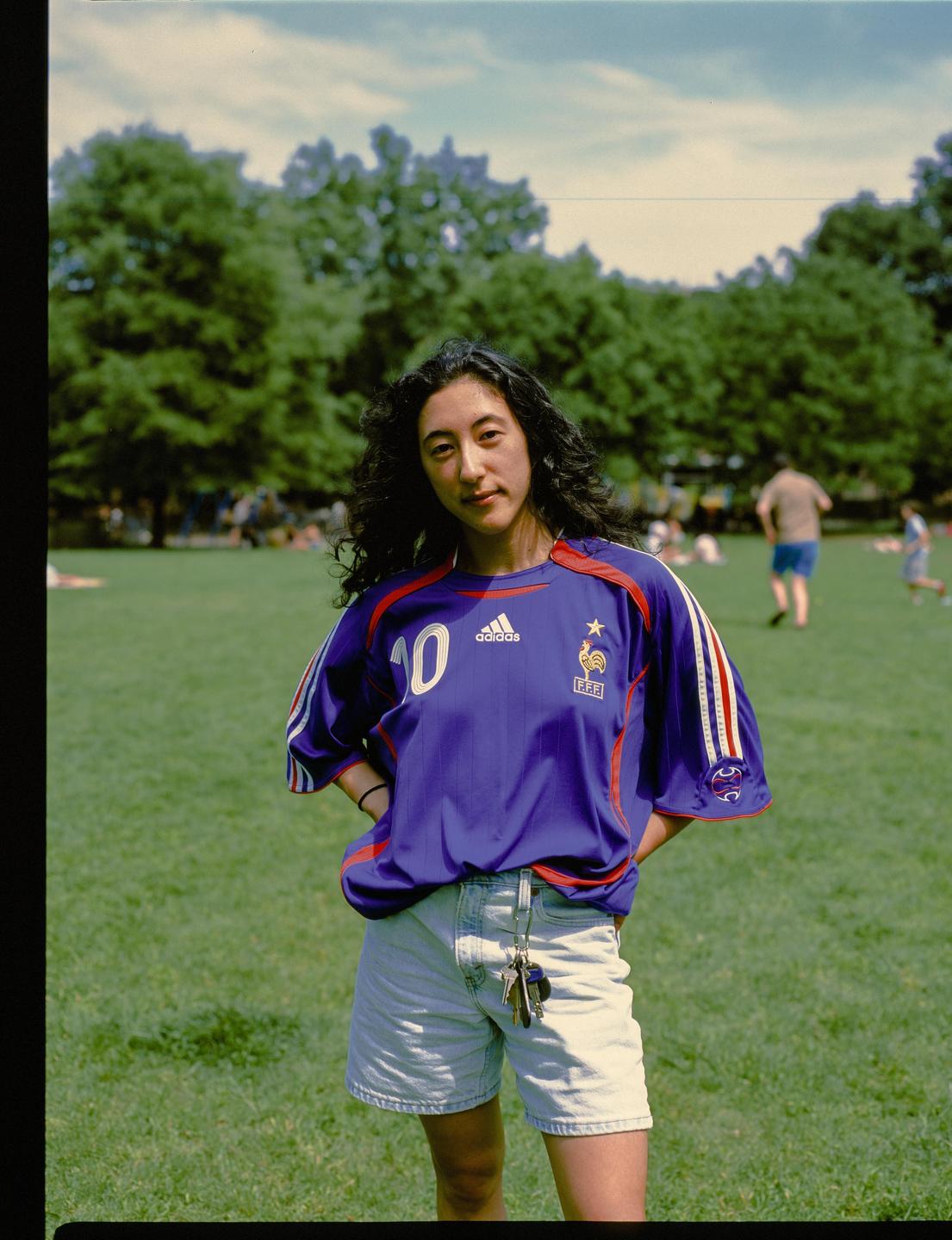 A woman wearing a blue soccer jersey and denim shorts stands in a grassy park.