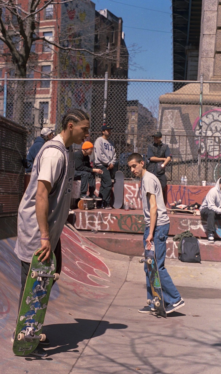 Two men hold skateboards in a skatepark with other people and buildings in the background.