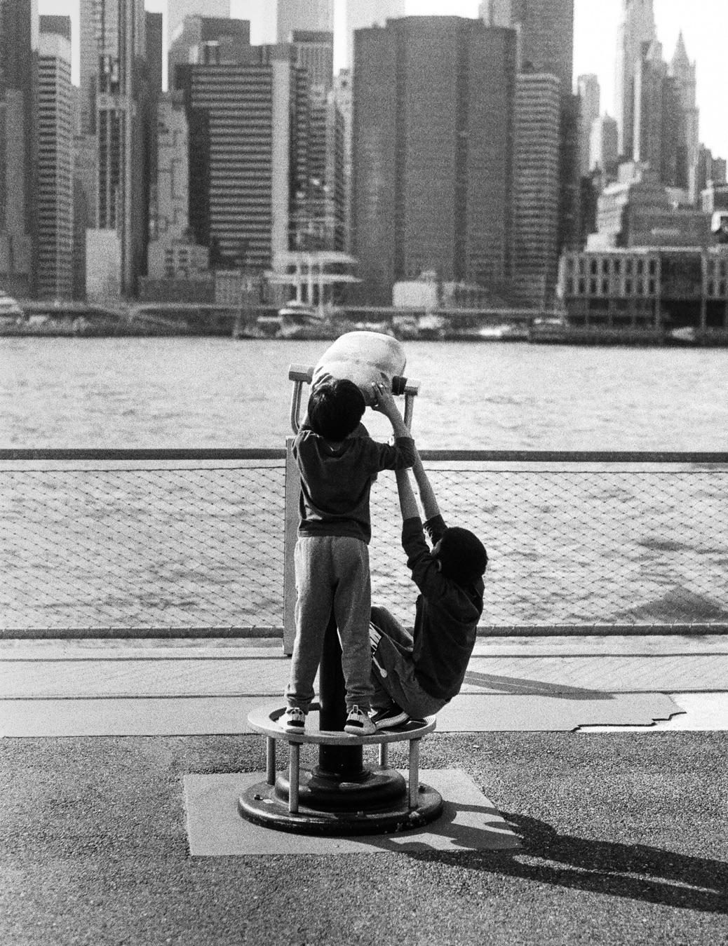 Two children play on a spinning ride with a city skyline and water in the background.