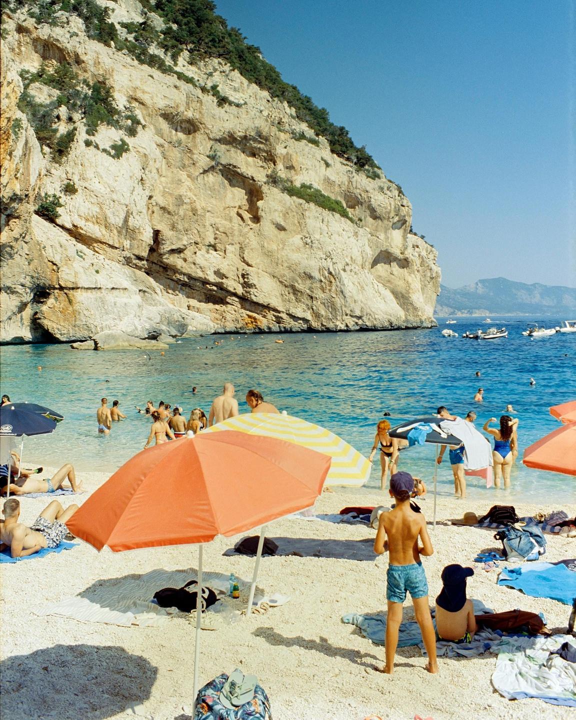 A beach with many people, colorful umbrellas, and boats in the water is nestled at the base of a large cliff.