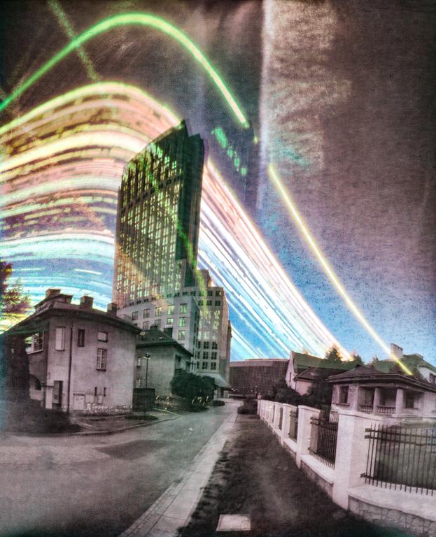 A street with houses and a tall building is illuminated by colorful light trails in the sky.