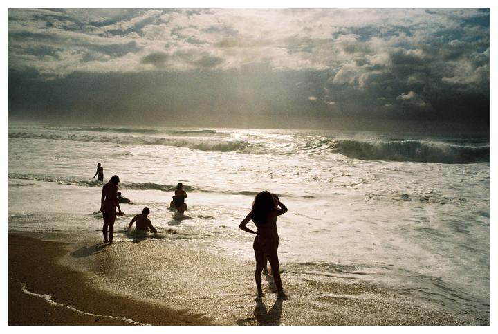 A group of people are silhouetted against the ocean waves and a cloudy sky at the beach.