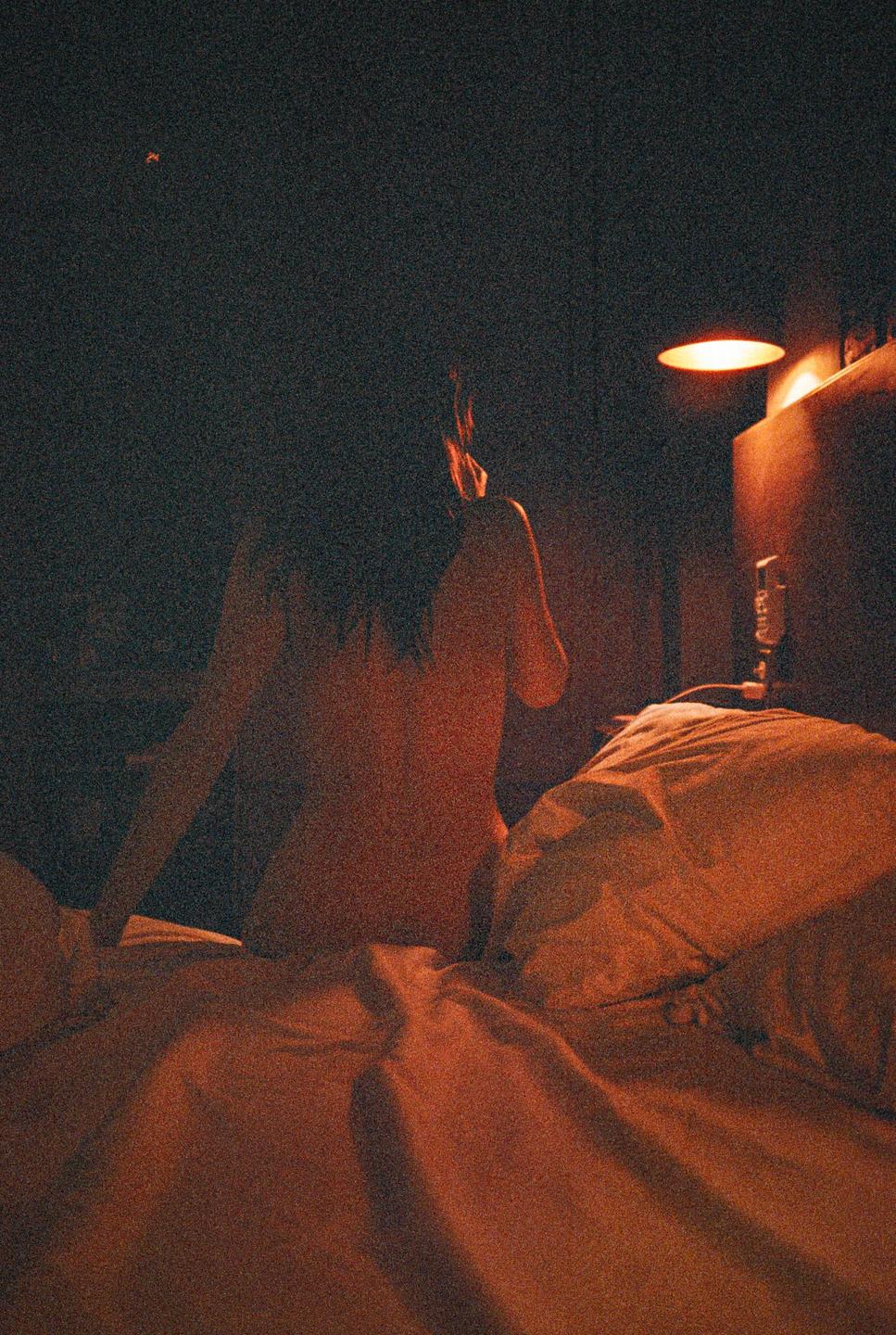 A woman sits on a bed in a dimly lit room with a lamp providing warm light.