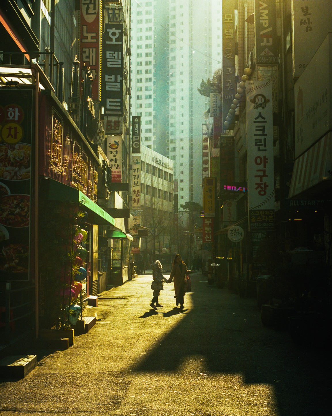 Two women walk hand in hand down a narrow street lined with tall buildings and signs in an urban setting.