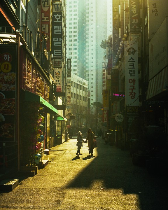 Two women walk hand in hand down a narrow street lined with tall buildings and signs in an urban setting.