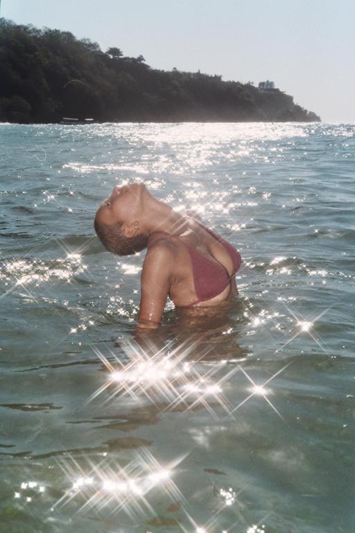 A woman with short hair and a bikini top is in the ocean with her head tilted back, surrounded by sparkling water.