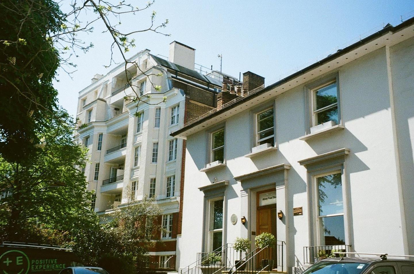 A white building with a brown door and a sign that reads "Abbey Road Studios" is visible next to a taller white building with balconies.