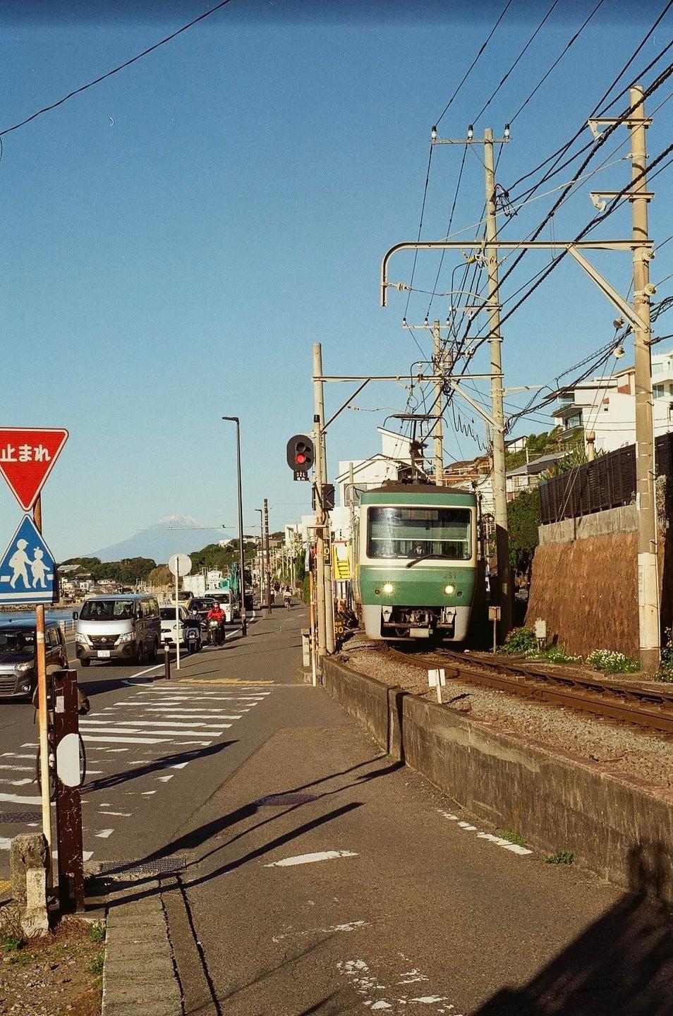 A green train is on tracks next to a street with cars and a mountain in the distance under a clear sky.