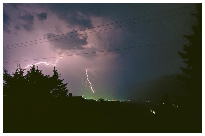 Lightning strikes over a distant town in the mountains at night, with silhouetted trees and power lines in the foreground.
