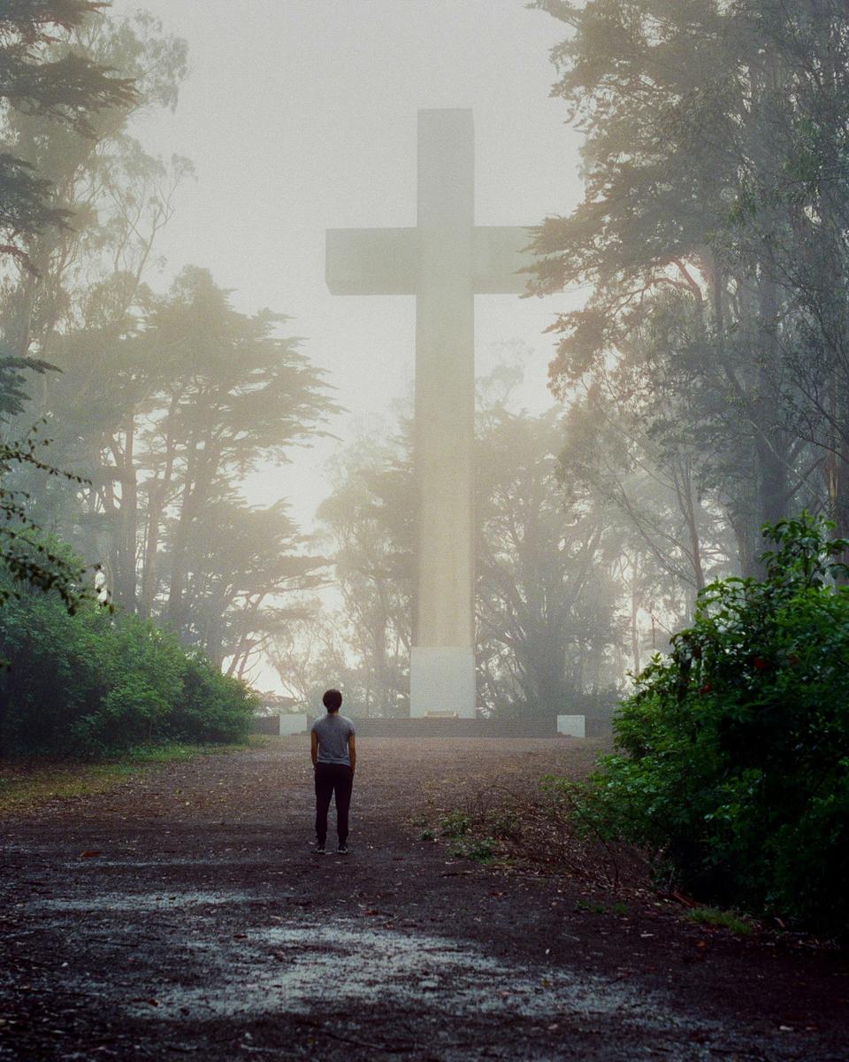 A person stands on a dirt path looking at a large cross in a hazy forest.