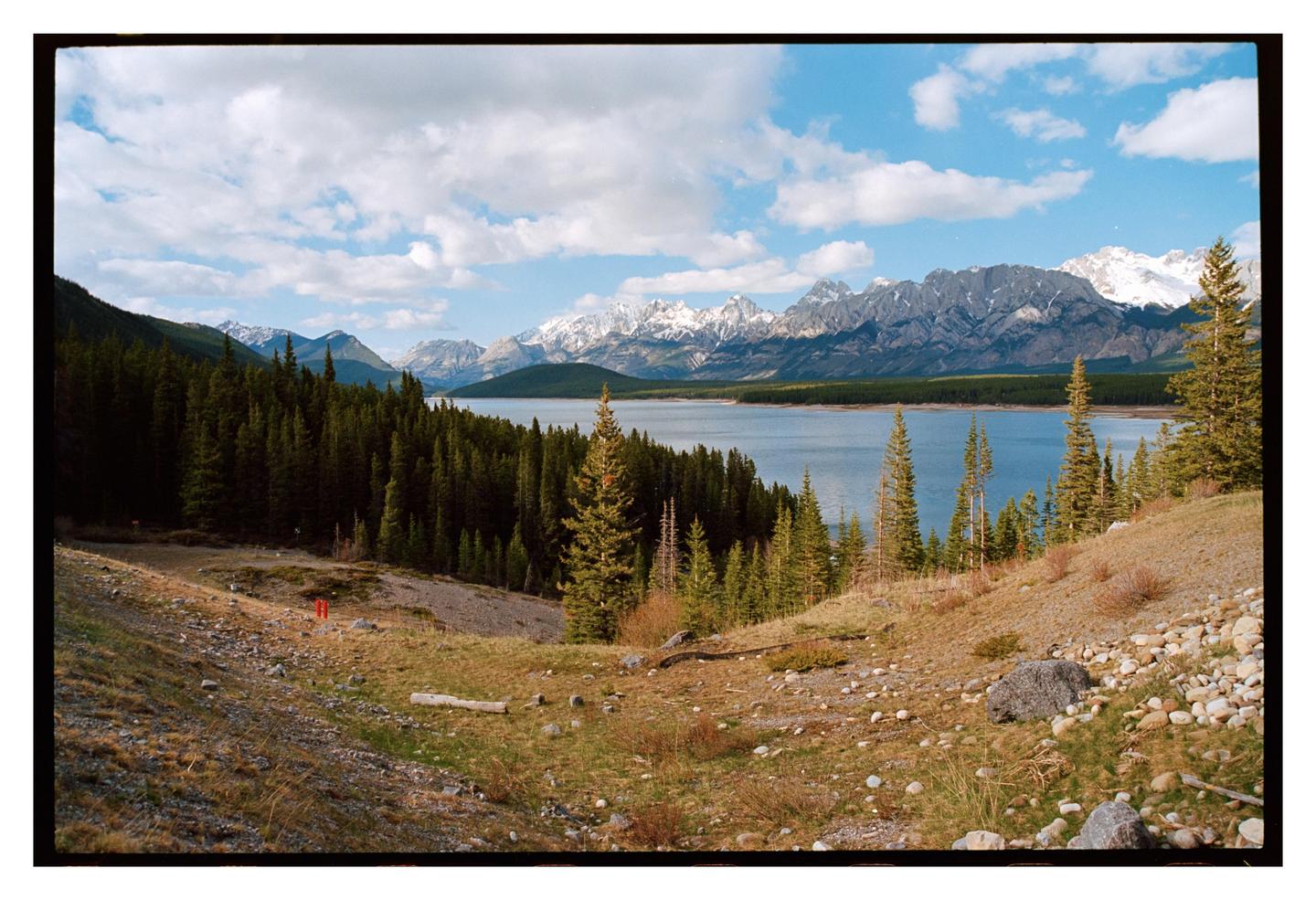 A wide shot shows a lake surrounded by a forest and snow-capped mountains under a cloudy sky.