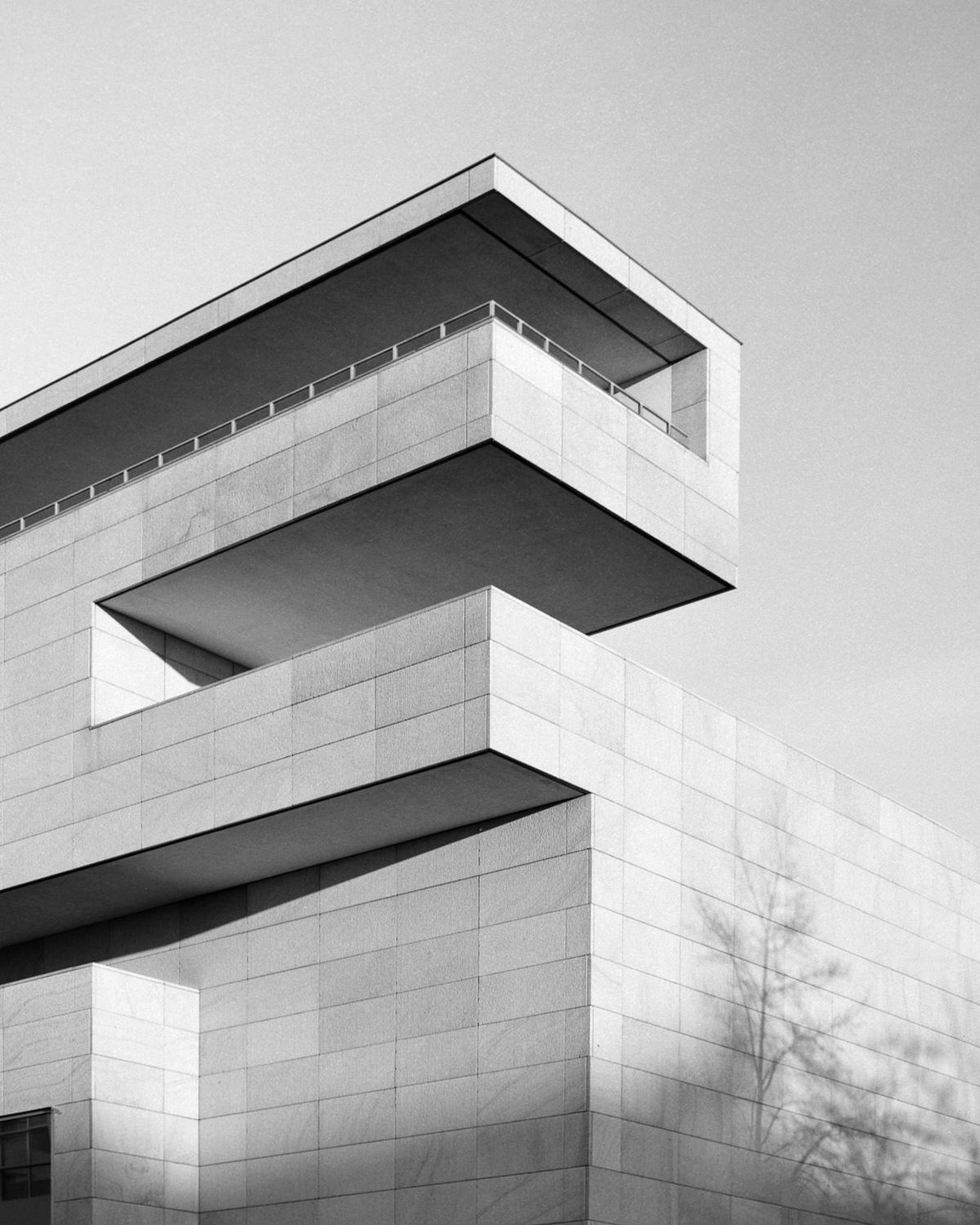 A modern building with multiple cantilevered sections and a tree shadow on its facade is shown from a low angle.