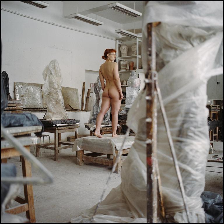 A nude woman stands on a wooden platform in an art studio filled with sculptures covered in plastic.