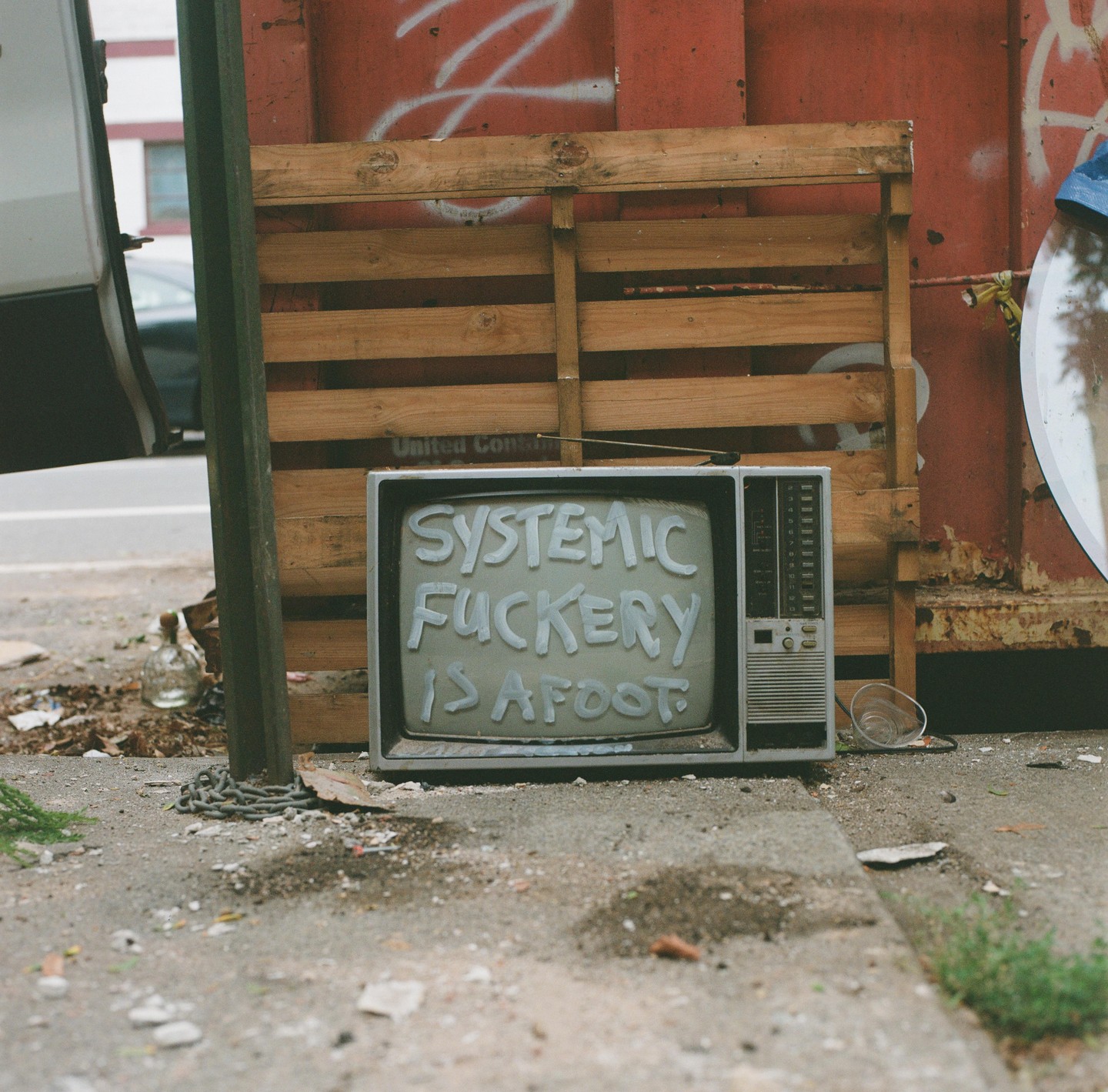 An old television with the words "SYSTEMIC FUCKERY IS AFOOT" painted on its screen sits on a sidewalk next to a wooden pallet and a red dumpster.