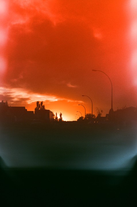 A vibrant orange sky with clouds hangs over a silhouetted street with streetlights and buildings.