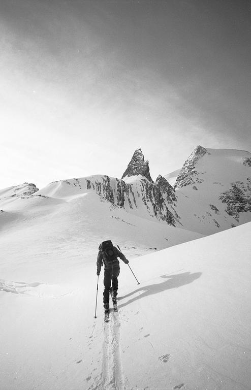 A person skis up a snowy mountain with a large backpack and ski poles.