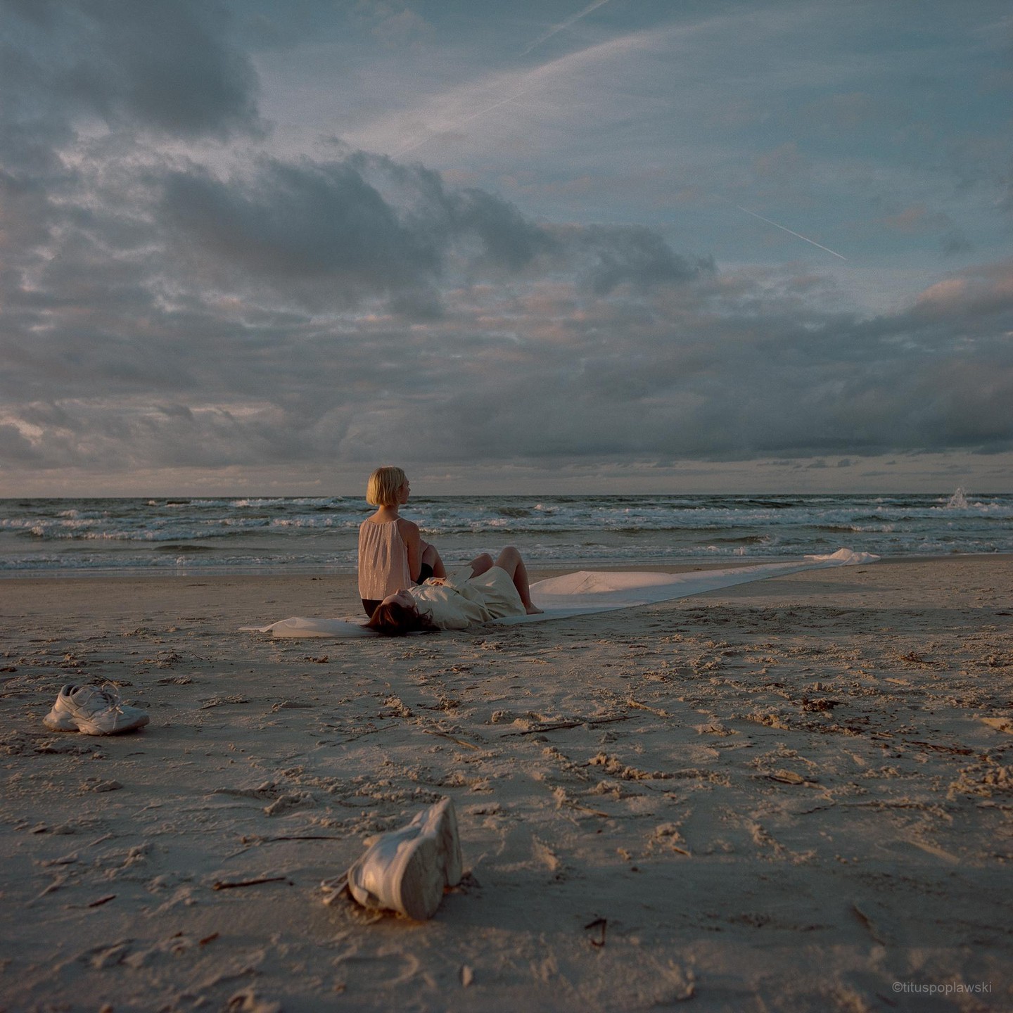 Two women are sitting on a blanket on a sandy beach looking out at the ocean under a cloudy sky.