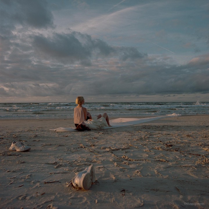 Two women are sitting on a blanket on a sandy beach looking out at the ocean under a cloudy sky.
