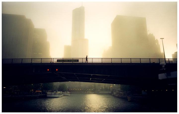 A person walks across a bridge over a river with city buildings in the hazy background.