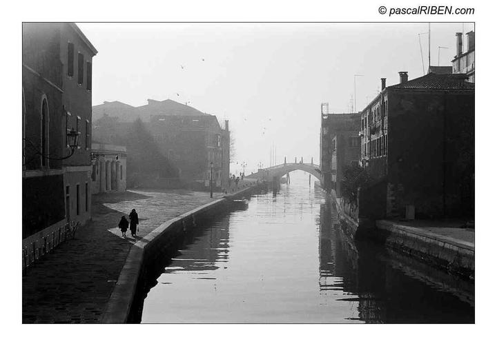 Two people walk along a canal with buildings on either side and a bridge in the distance.