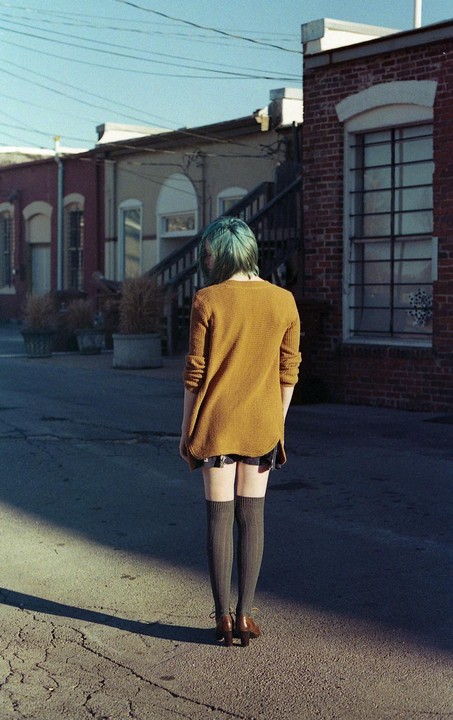 A woman with blue hair and a mustard yellow sweater stands in an alley with brick buildings.