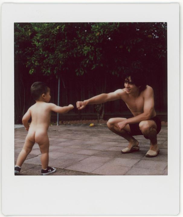 A man and a child are fist-bumping on a paved patio in front of a green bush.