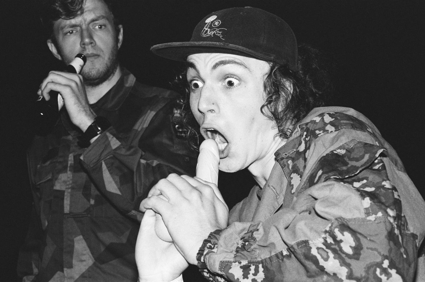 A man in a camouflage jacket and baseball cap holds a white object to his open mouth while another man drinks from a bottle in the background.
