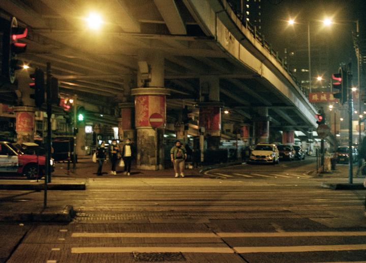 A street at night is illuminated by streetlights and traffic lights, with an overpass above and cars and people below.