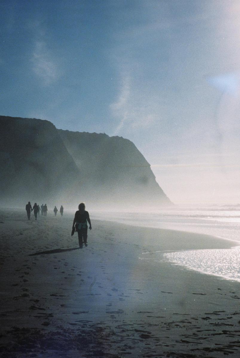A group of people walk along a hazy beach with cliffs in the background.