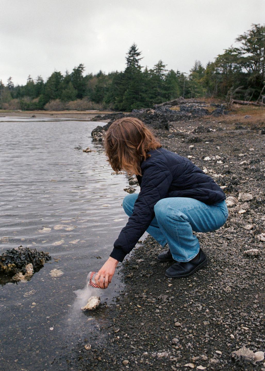 A woman crouches by the water's edge, pouring ashes from a small container onto a rock.