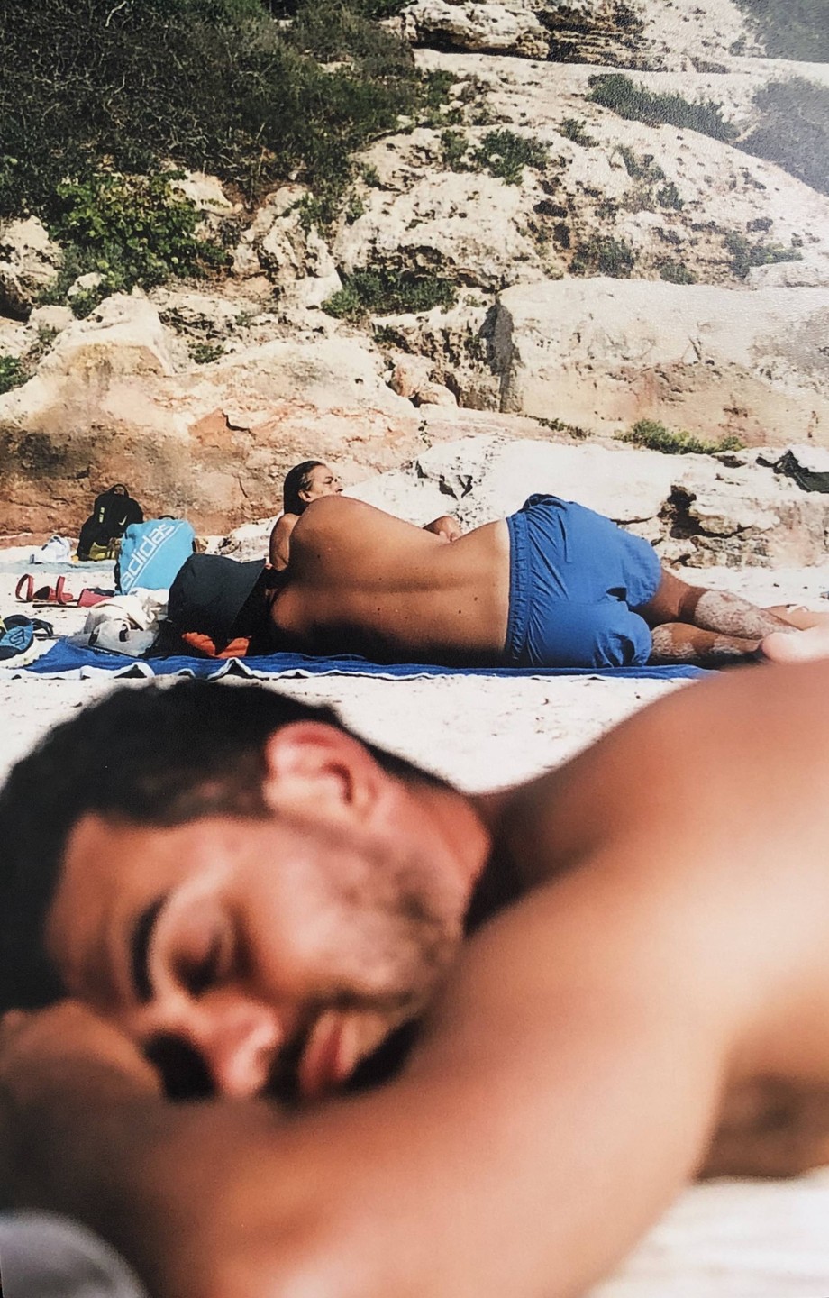 Two men are napping on a sandy beach with rocky cliffs in the background.