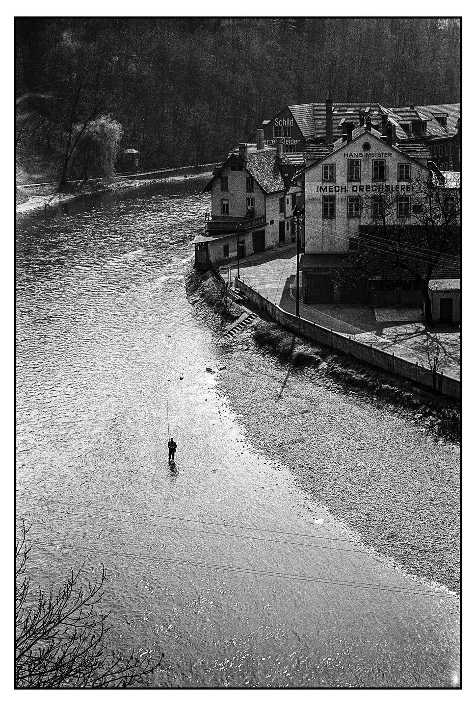 A lone fisherman stands in a river next to a town with buildings and trees.