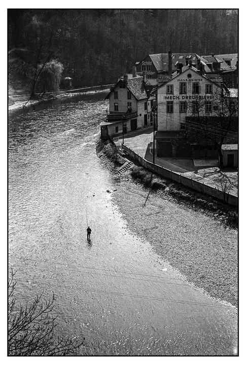 A lone fisherman stands in a river next to a town with buildings and trees.