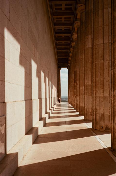 A person sits at the end of a long hallway lined with columns and shadows.