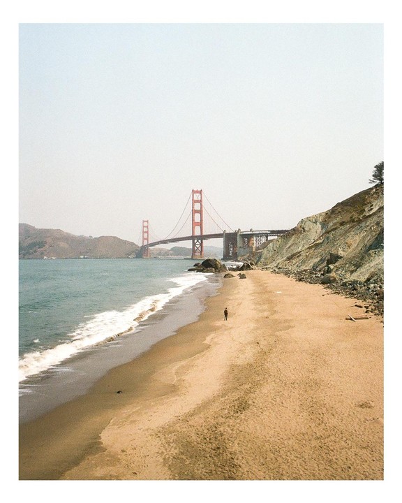 A person walks on a sandy beach with waves crashing on the shore and the Golden Gate Bridge in the background.