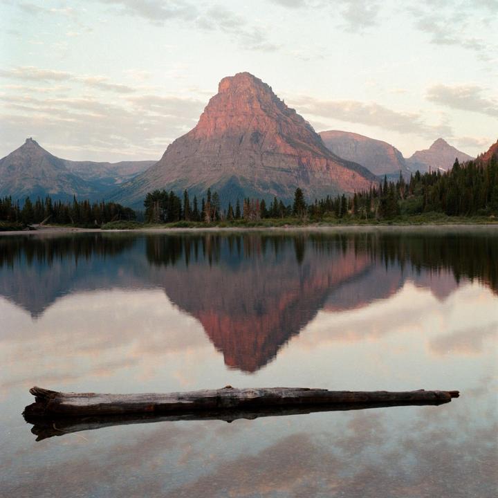 A large mountain reflects in a still lake with a log floating in the foreground.