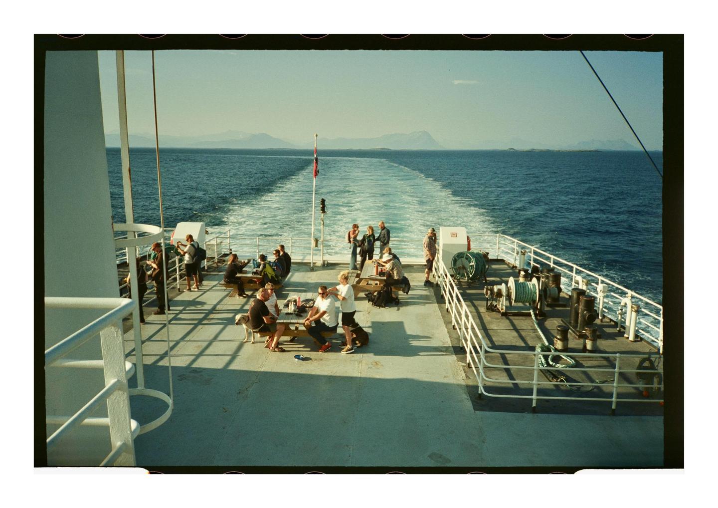 People are gathered on the deck of a ferry, leaving a white wake behind it on the blue water.