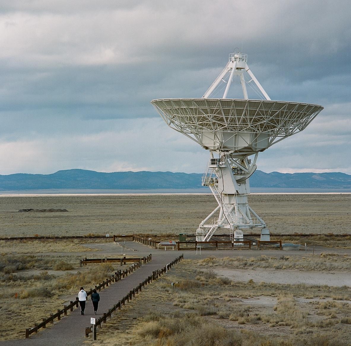 Two people walk on a path towards a large white radio telescope under a cloudy sky.