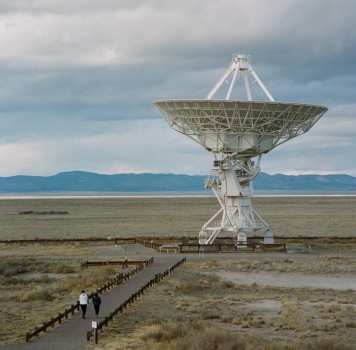 Two people walk on a path towards a large white radio telescope under a cloudy sky.