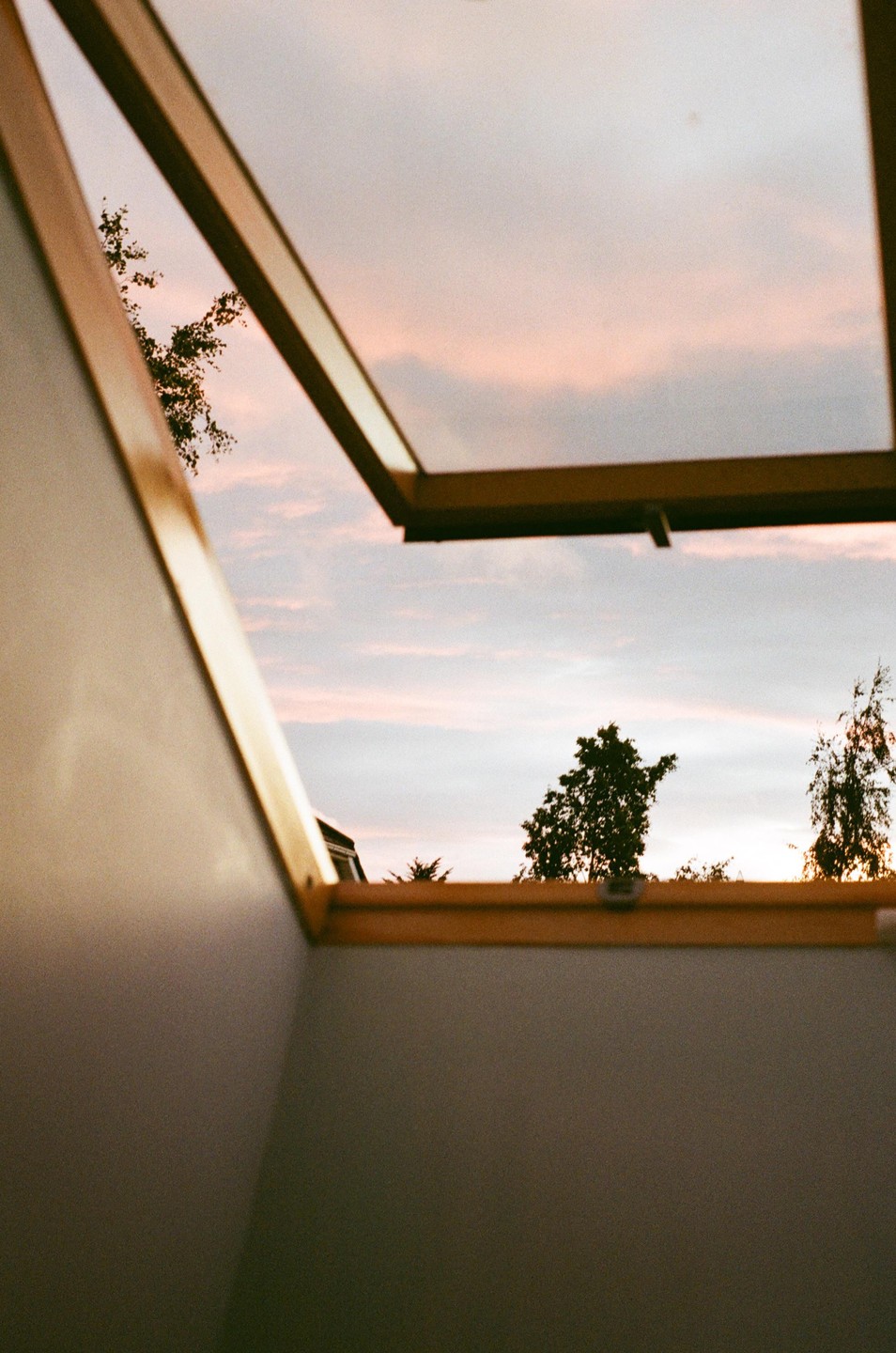 A view of a pink and blue sunset sky with trees and a house through an open skylight.
