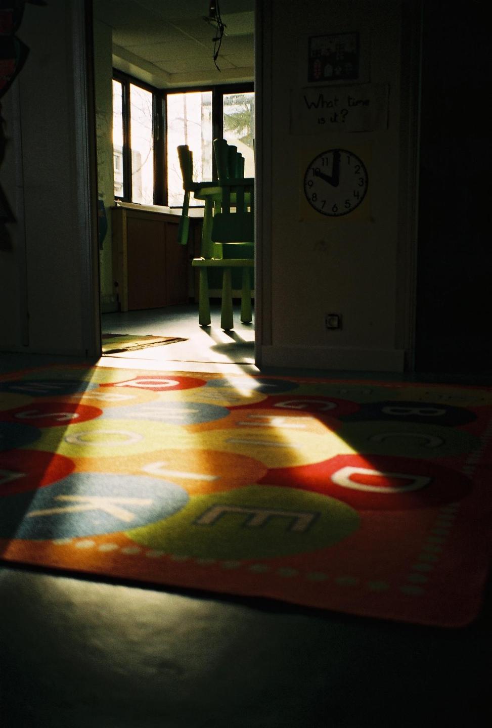Sunlight illuminates a colorful alphabet rug in a room with green chairs and a clock on the wall.