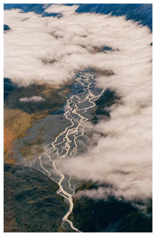 An aerial view shows a braided river winding through a mountainous landscape partially obscured by clouds.