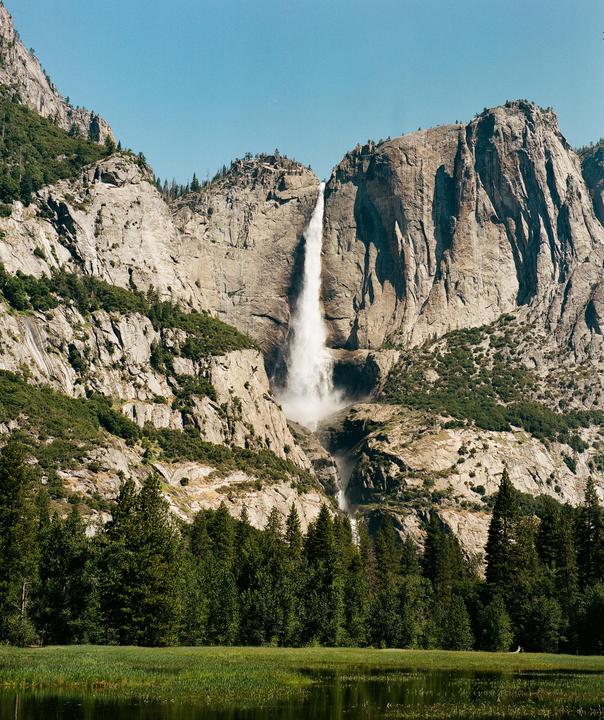 A tall waterfall cascades down a rocky mountain face into a forest and a body of water.