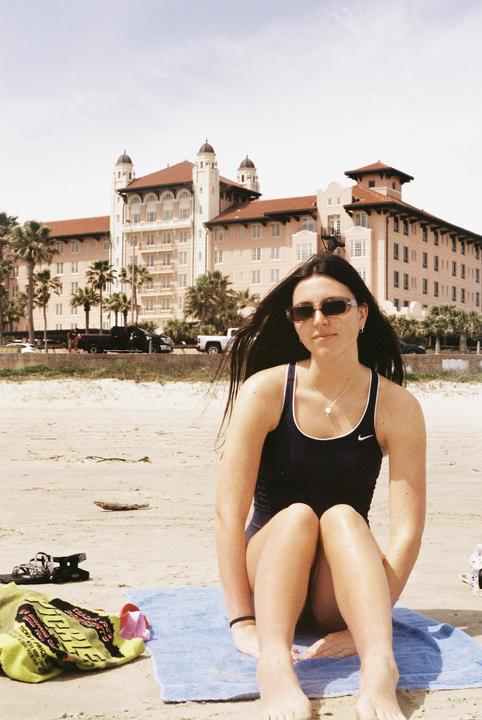 A woman in a swimsuit sits on a towel on a sandy beach with a large building and palm trees in the background.