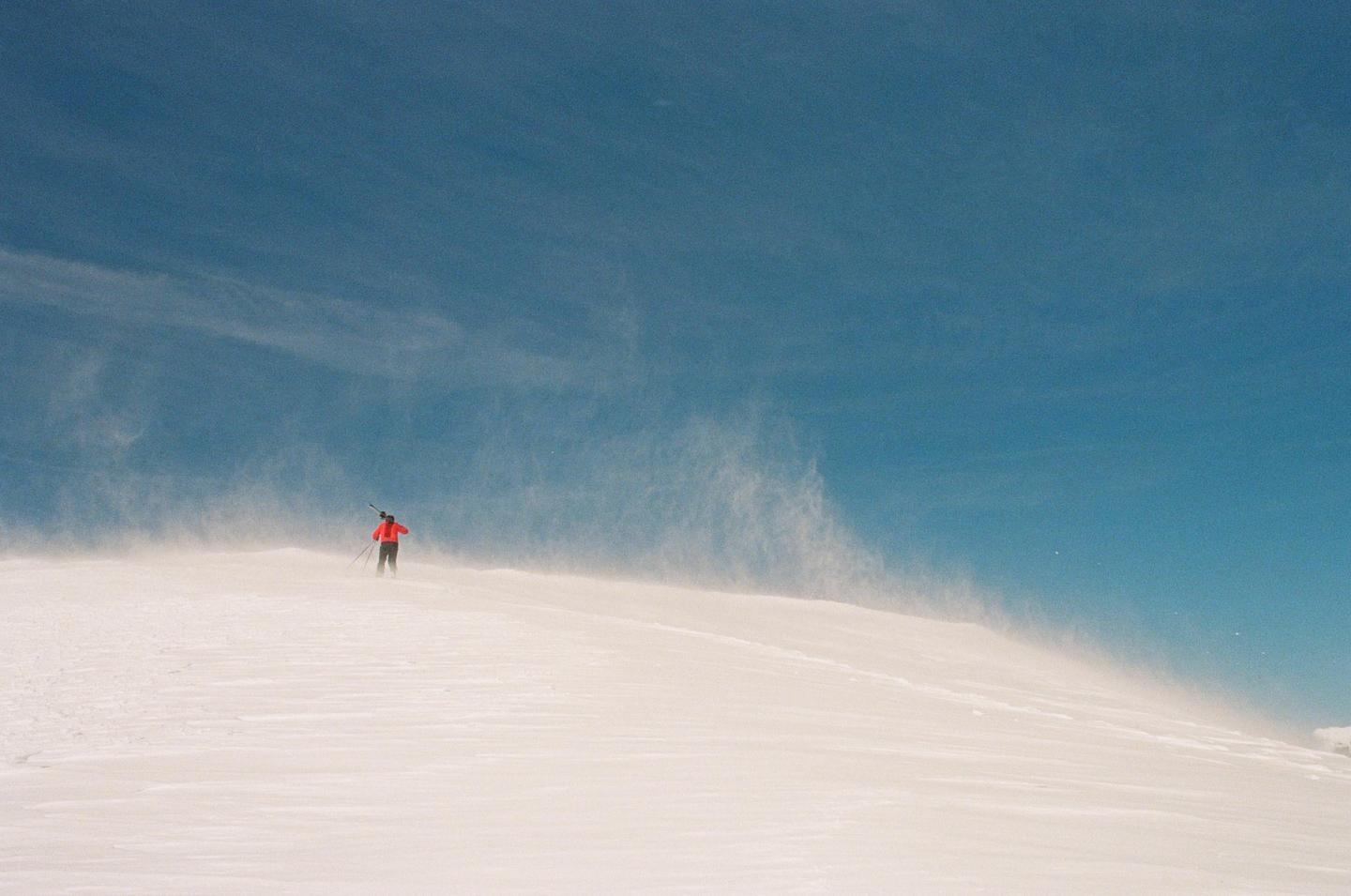 A person in a red jacket stands on a snow-covered hill with snow blowing around them under a clear blue sky.