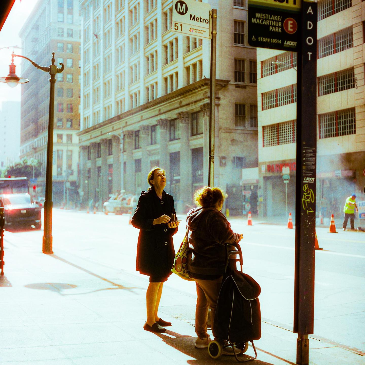 Two women stand on a sunlit street corner next to a Metro bus stop sign, with tall buildings and a street lamp in the background.