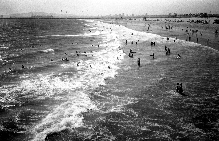 A large number of people are swimming and wading in the ocean near a beach with a city skyline in the distance.