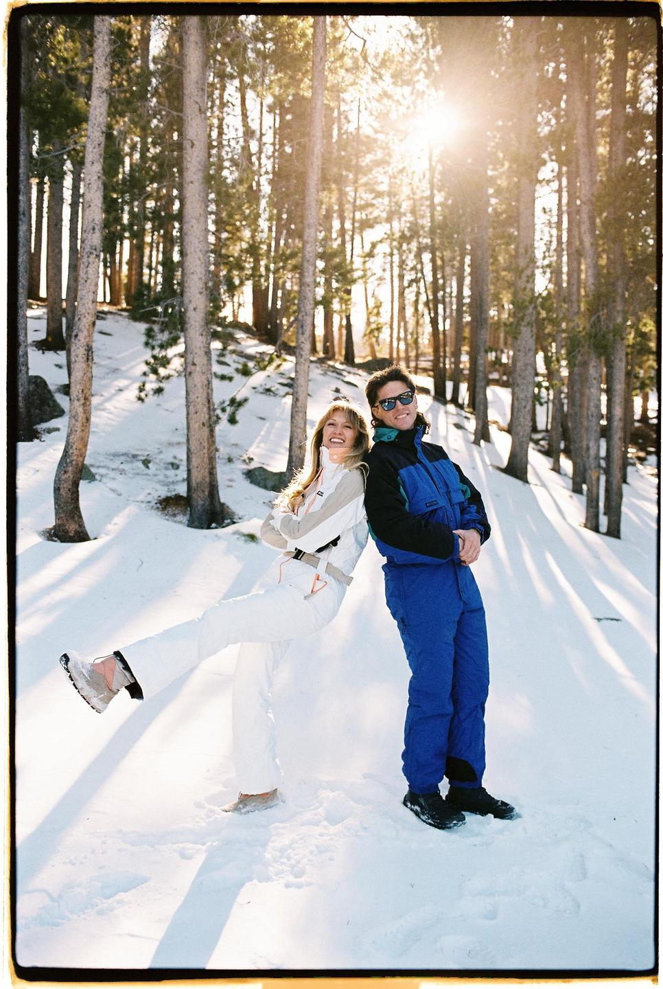 A couple in ski suits stands back-to-back in a snowy forest with sunlight streaming through the trees.